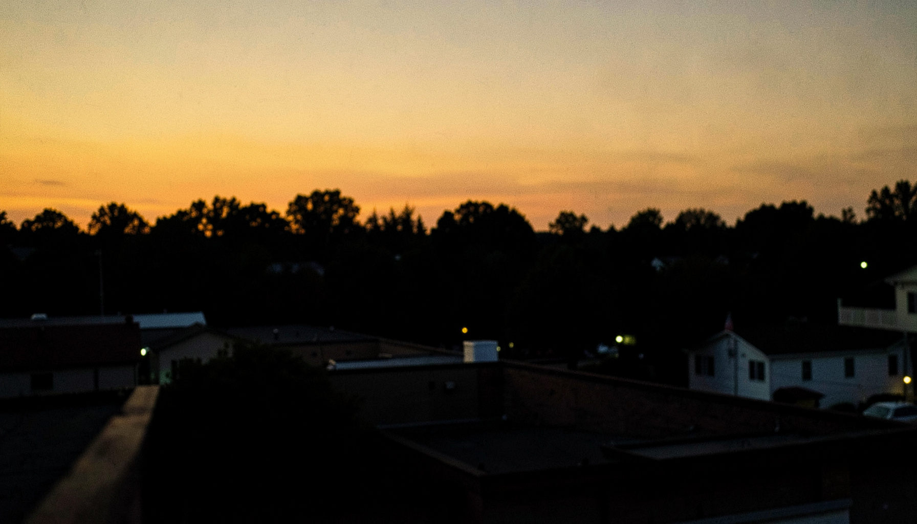 Atmospheric dusk view across Fort Collins rooftops with warm amber sky against deep near-black silhouette of trees and hills